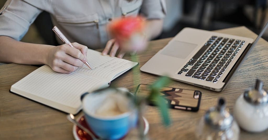 Woman writing in a notebook with a laptop and coffee cup on a desk. Ideal for workspace inspiration.