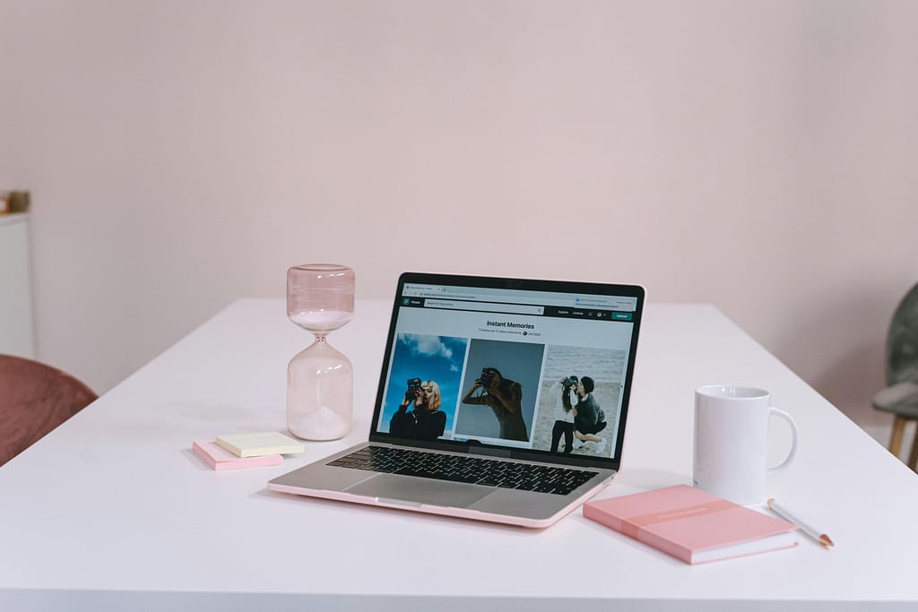 A clean and minimalist workspace featuring a laptop, hourglass, notebook, and coffee mug on a white desk.
