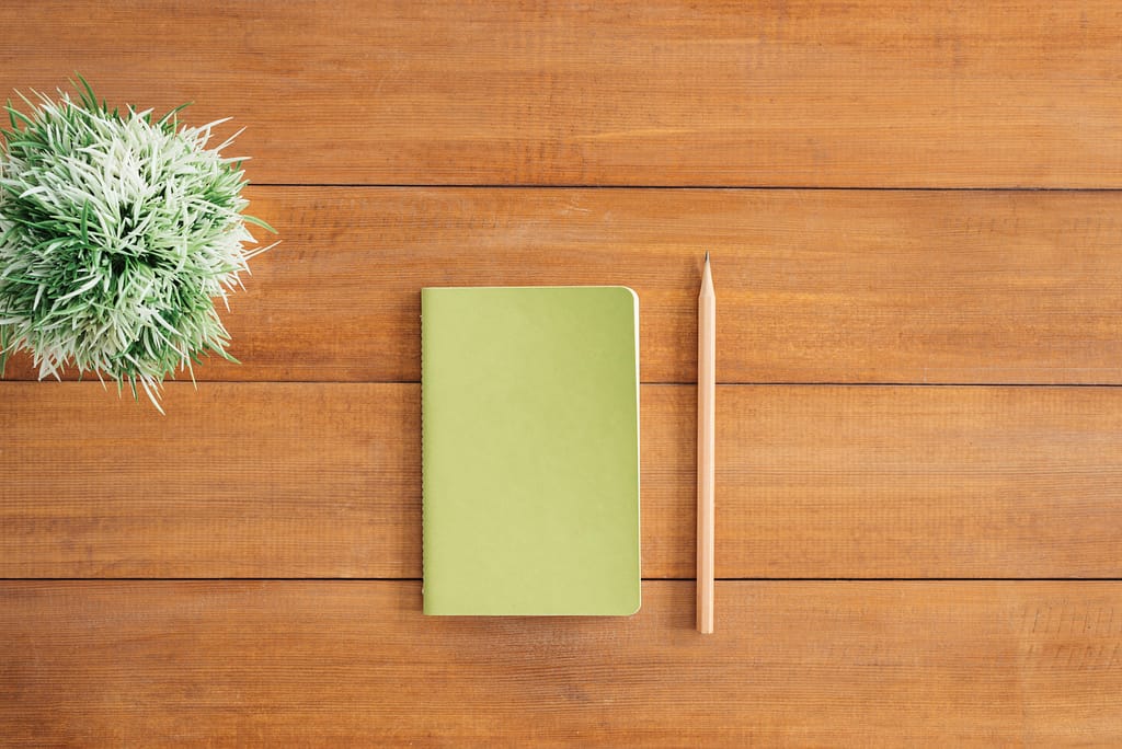 Flat lay of green notebook and pencil on wooden desk with plant. Perfect for creativity and organization themes.