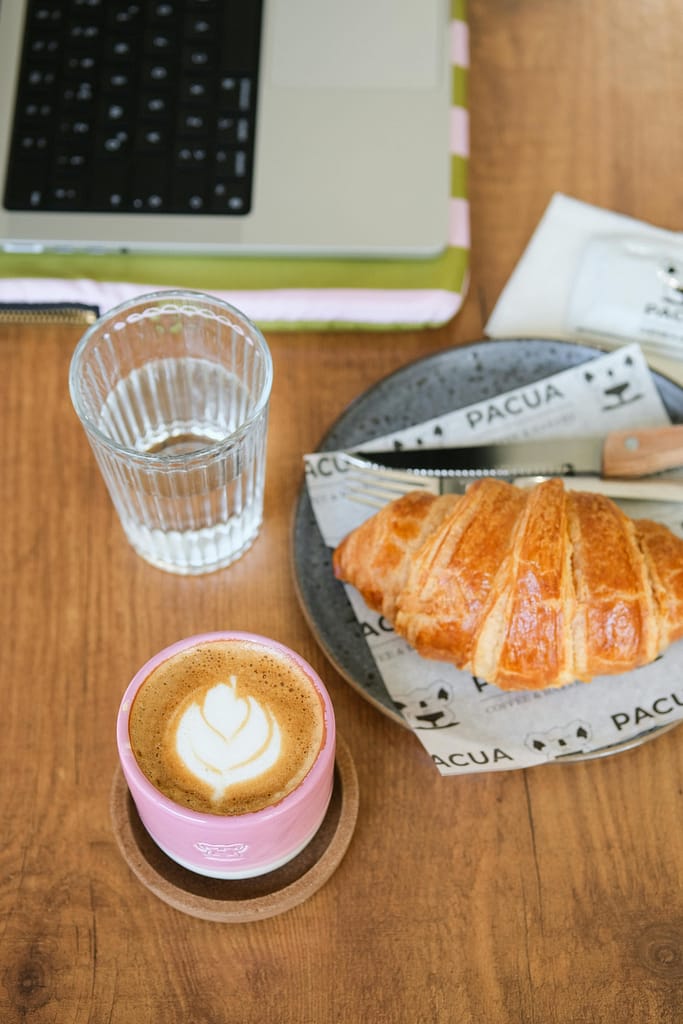 A cozy morning scene with coffee and croissant on a wooden table next to a laptop.