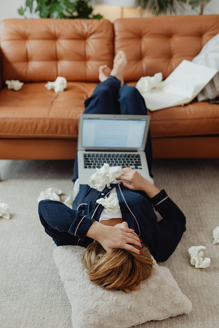 Woman in loungewear, stressed, lies on floor with laptop amidst crumpled papers indoors.